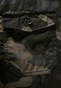 Maskovacia tkanina s odtieňmi tmavo zelenej a hnedej, s náprsným vreckom a čiernym gombíkom. Hladká textúra a prešité detaily.