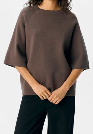 Femme portant un pull marron à manches courtes ample et un pantalon noir, debout les mains doucement jointes devant elle.