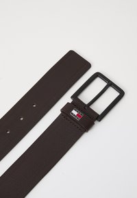 Cintura in pelle marrone con finitura testurizzata, dotata di una fibbia in metallo nero e un dettaglio con logo in rosso, bianco e blu sul cinturino.
