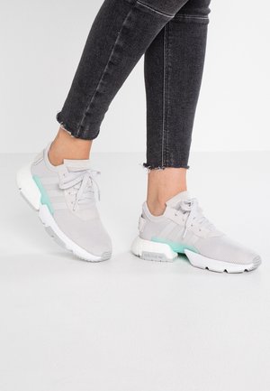 Personne portant des baskets Adidas gris clair avec des semelles blanches et des accents turquoise, associées à un jean skinny noir à ourlet effiloché.