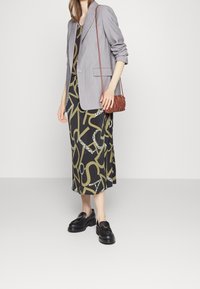 Grau Blazer aus glattem Stoff, schwarzes Kleid mit grünem abstraktem Muster, braune Handtasche und schwarze Mokassins mit dicker Sohle.