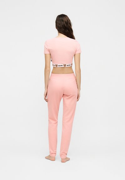 Γυναίκα που στέκεται ξυπόλυτη, φοράει ροζ cropped T-shirt και ροζ παντελόνια που ταιριάζουν με διακόσμηση Moschino στη μέση, με την πλάτη στραμμένη.