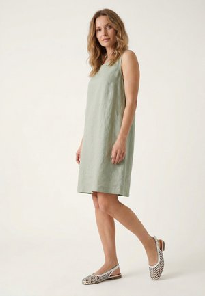 Femme aux cheveux ondulés portant une robe sans manches vert clair longueur genou et des chaussures plates blanches tissées, debout sur un fond clair uni.