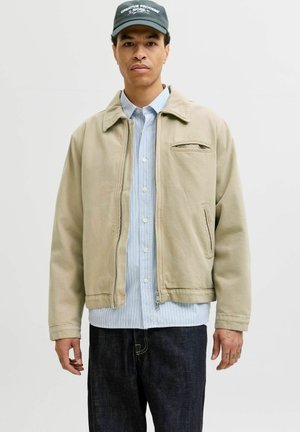 Moški v bež jopiči s zadrgo čez modro srajco s črtami in temnih kavbojkah, z rjavo kapo z napisom "Creative Process Work".