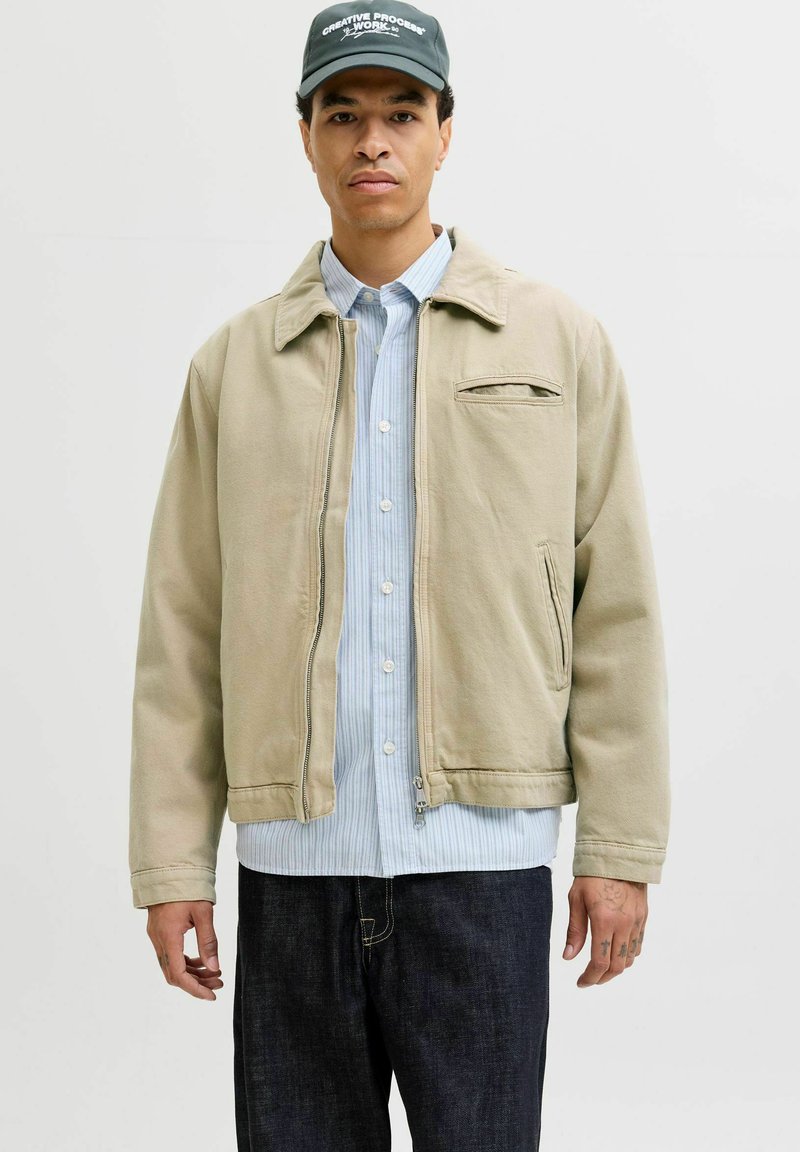 Mann trägt eine beige Reißverschlussjacke über einem blau gestreiften Hemd und dunkle Jeans, mit schwarzer Mütze mit der Aufschrift "Creative Process Work."