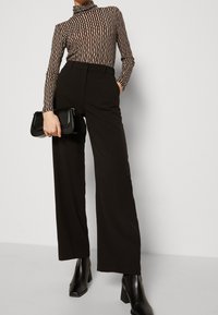 Svarta vida byxor i kombination med en mönstrad långärmad topp med vertikala ränder i beige och mörkbrun, samt en svart kuvertväska.