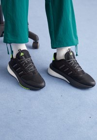 Zapatillas deportivas negras con parte superior de malla, cordones grises y acentos en verde neón. Suela blanca gruesa diseñada para la comodidad. Llevadas con calcetas blancas.
