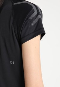 Schwarzes, kurzärmeliges Sportshirt mit strukturierten grauen Akzenten an den Ärmeln und einem "D1"-Etikett. Das Material wirkt glatt und dehnbar.