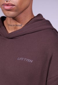 Sudadera marrón hecha de tejido suave, con un bolsillo canguro y un logo "URTRN" en gris claro en la parte izquierda del pecho.