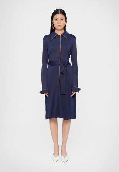 Marineblauwe jurk met een knoopsluiting aan de voorkant, lange mouwen, oranje biezen en een ceintuur om zelf te strikken. Witte puntige schoenen maken de look af.