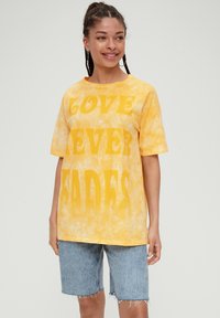 Żółta koszulka tie-dye z wyraźnym białym napisem "MIŁOŚĆ NIGDY NIE ZNIKA", okrągły dekolt i krótkie rękawy. Połączona z dżinsowymi szortami.