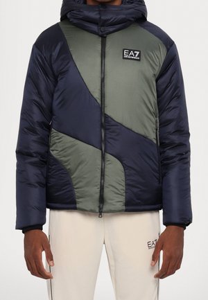 Homme portant une veste matelassée bleu marine et vert olive avec fermeture éclair à l’avant et logo EA7 Emporio Armani sur la poitrine, ainsi qu’un pantalon beige avec logo.