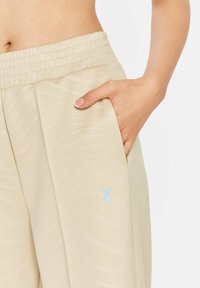 Beige, lichte joggingbroek met een soepele textuur, elastische tailleband, zijzakken en een klein blauw logo op de onderrand.