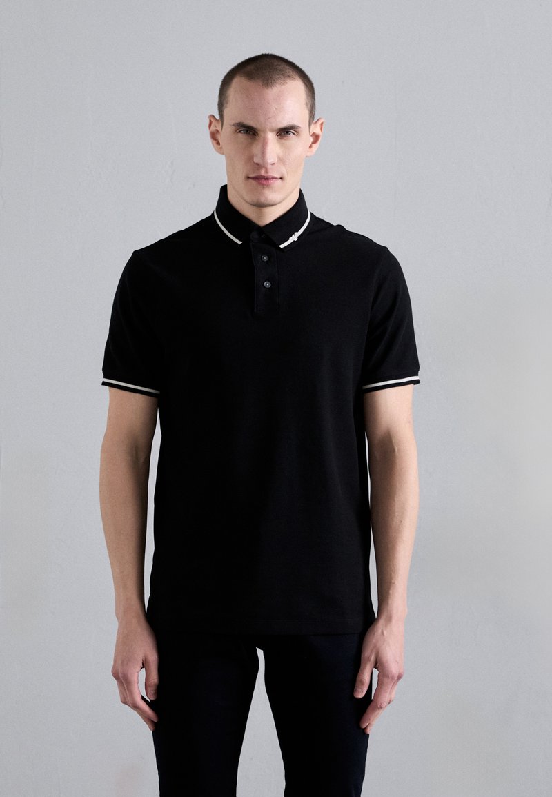Black polo shirt made of cotton, featuring a collared neck with white accents and a four-button placket. Short sleeves with striped cuffs.