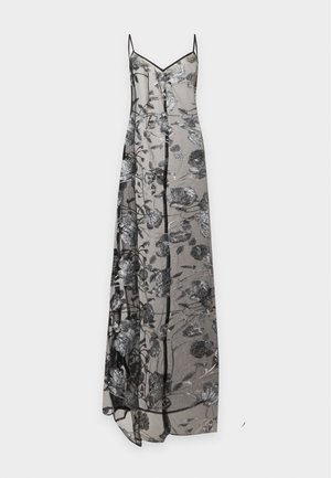 Femme portant une robe maxi noire transparente avec une broderie florale argentée par-dessus un soutien-gorge et des sous-vêtements noirs, debout avec une main sur la hanche.