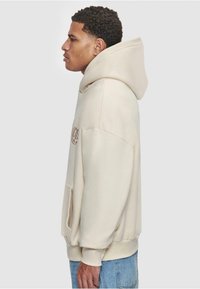 Haut à capuche beige avec une grande capuche, poche frontale et poignets côtelés. Présente un logo brodé sur la poitrine. Coupe décontractée avec une texture douce.