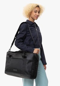 Musta viestintä laukku, jossa on teksturoitu kangas, kaksoisetteinen yläkäsin, irrotettava olkahihna ja etupuolella vetoketjullinen tasku.