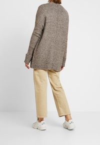 Brązowy sweter z dzianiny z prążkowanym dołem. Połączony z beżowymi spodniami w szerokim kroju i białymi trampkami. Sweter ma luźny krój i detal w postaci szwu z tyłu.