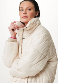 Chaqueta puffer crema con un cuello alto, cierre de cremallera y textura acolchada diagonal. Los puños presentan un detalle en contrastante beige y rosa.