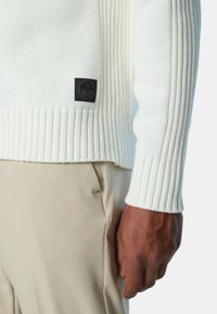 Maglione lavorato a maglia color crema con texture a coste, dotato di un patch logo nero all'orlo. Indossato sopra pantaloni beige, con un primo piano del maniche e della mano.