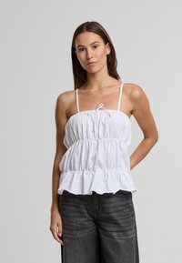 Biely bezrukávový top s nastaviteľnými ramienkami, nariasenou prednou časťou a volánovým spodným okrajom. V kombinácii s tmavosivými širokými nohavicami.