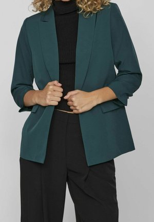 Femme portant un blazer vert foncé sur un pull à col roulé noir et un pantalon noir taille haute, tenant les revers du blazer avec les deux mains.