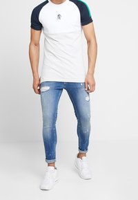Camiseta blanca con mangas raglán azul marino y acentos verdes, combinada con jeans skinny desgastados azul claro y zapatillas blancas.