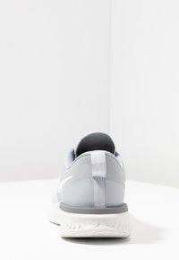 Zapatilla deportiva gris con una parte superior de tela texturizada, cuello acolchado y suela de goma blanca. Presenta un logotipo en el lateral y una tira de agarre en el talón.