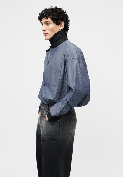 Άνδρας με σγουρά μαλλιά που φοράει γαλάζιο πουκάμισο με λεπτές κάθετες ρίγες, υψηλό μαύρο γιακά και ξεθωριασμένα μαύρα τζιν, στέκεται σε πλάγια θέση με τα χέρια στις τσέπες.