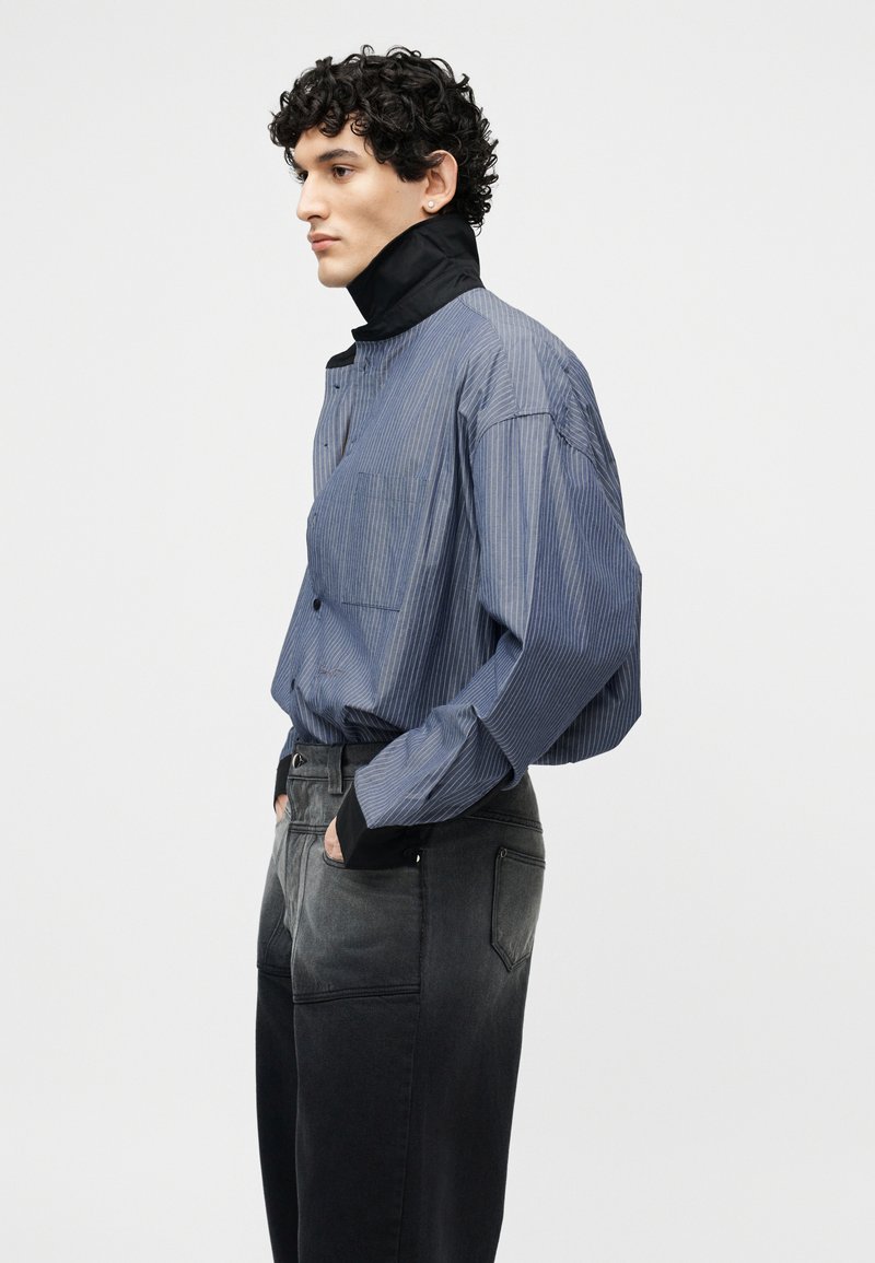 Man with curly hair wearing a blue pinstripe shirt with a high black collar and faded black jeans, standing in profile with hands in pockets.