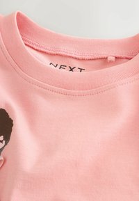 Tricou din bumbac roz deschis, cu guler rotund, mâneci scurte și un design grafic negru pe față. Eticheta brandului este vizibilă în interiorul gulerului.