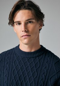 Dunkelblauer Strickpullover mit Zopfmuster, Rundhalsausschnitt und strukturiertem Stoff. Das Model hat kurzes, leicht welliges Haar.