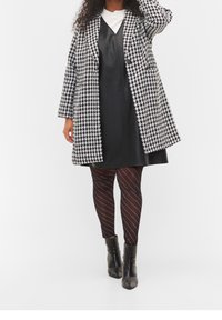 Abrigo de pata de gallo en blanco y negro sobre un vestido de cuero negro, combinado con leggings de rayas negras y botas de tobillo negras.