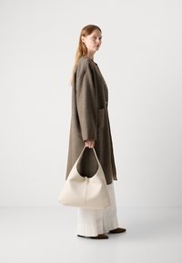 Brun ullkåpe med to knapper og lommer, sammen med en kremfarget hobo-veske med et rundt håndtak og minimalistisk design. Hvite bukser.