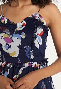 Robe florale bleu marine avec bretelles et ourlet à volants. Présente des motifs floraux colorés en rouge, jaune, bleu et blanc sur un tissu léger.