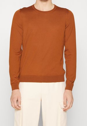 Uomo che indossa un maglione aderente a maniche lunghe color ruggine e pantaloni cargo beige chiaro, in piedi con le braccia rilassate ai lati.