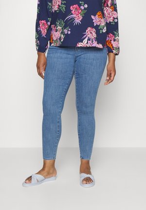 Frau trägt hellblaue enge Jeans, lila Bluse mit Blumenmuster und langen Ärmeln sowie hellblaue Sandalen mit offenem Zehenbereich und steht auf einem schlichten weißen Boden.