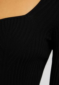 Haut en maille côtelée noire avec un décolleté asymétrique dévoilant une épaule sur une personne à la peau claire.