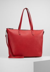 Červená kožená taška přes rameno s dvojitými ramenními popruhy, odnímatelným popruhem na kříž a horním zipem na bílém podkladu.