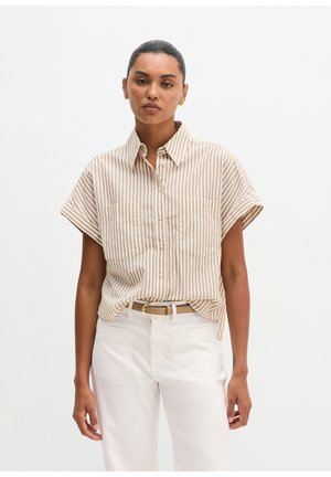 Woman wearing a beige and white vertical striped short-sleeve button-up shirt tucked into white pants with a tan belt.