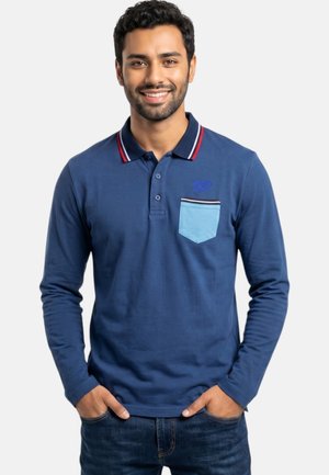 Joven con pelo corto y oscuro, vistiendo una camisa tipo polo azul de manga larga con un bolsillo azul claro en el pecho, sonriendo con las manos en los bolsillos de los vaqueros.