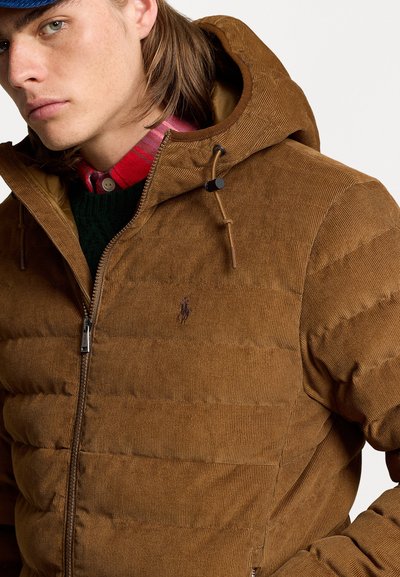 Brown corduroy puffer jacket with a zip front, hood, and toggle adjustments. Features a subtle logo on the chest and layered with a plaid shirt.