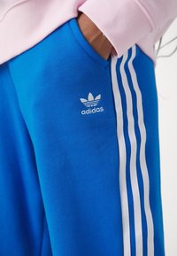 Pantaloni della tuta blu con strisce bianche lungo i lati. Presentano il logo Adidas bianco sulla coscia superiore. Realizzati in tessuto morbido e liscio.