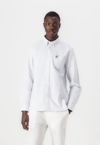 Chemise boutonnière bleu clair avec des rayures verticales blanches, poche poitrine et écusson logo jaune. Manches longues avec poignets boutonnés.