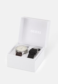 Montre argentée avec un cadran blanc et un bracelet en cuir marron, accompagnée d'un bracelet en silicone noir, présentée dans une boîte blanche avec le logo "GUESS".