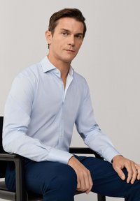 Muž v světle modré košili a tmavě modrých kalhotách sedí na černé židli před jednobarevným pozadím, čelem dopředu.