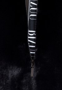 Zbliżenie częściowo rozpiętej czarnej futrzanej kurtki, pod którą widać materiał w czarno-białe napisy.