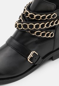 Botín negro de cuero hasta el tobillo con detalles de cadena dorada en capas y una correa con hebilla, con una textura suave y un diseño de punta redonda.