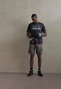 Homme portant des gants de boxe noirs, un T-shirt noir Venum et un short gris, se tenant contre un mur clair et regardant ses gants.