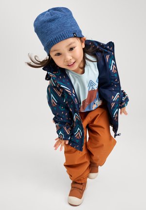 Colorful waterproof jacket with geometric patterns, orange pants, blue knit hat, and brown shoes. Child leaning forward with playful expression.
